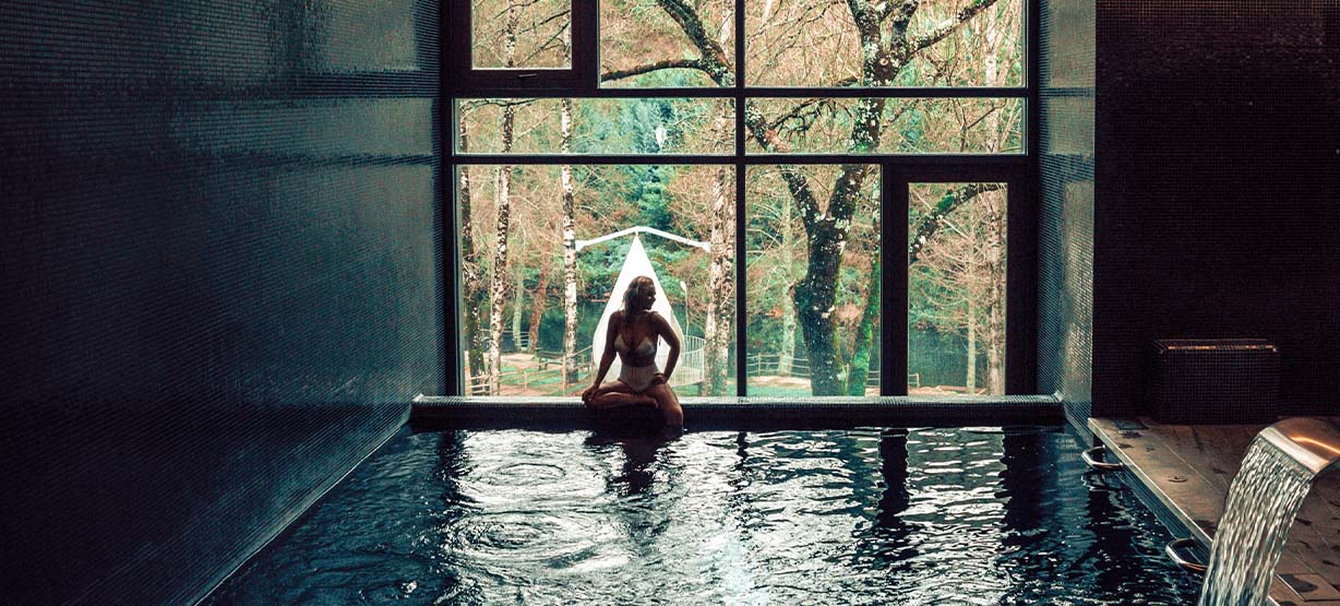 Mulher a posar em contraluz numa piscina termal, sentada perto de uma enorme janela no meio da natureza num hotel